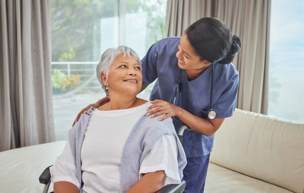 Female caregiver standing behind seated senior woman with her arms on her shoulders