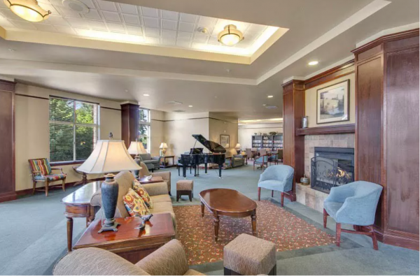 The common area at Merrill Gardens at Tacoma, with seating in front of a fireplace and a grand piano and the library area in the background