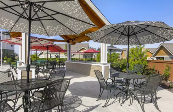 Multiple wrought iron tables and chairs, all shaded by umbrellas, on the patio at Merrill Gardens at Tacoma