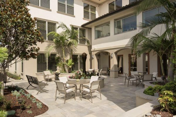 Outdoor patio in the corner of two buildings with outdoor tables and chairs and beautiful landscaping of trees and flowers at Oakmont of Pacific Beach