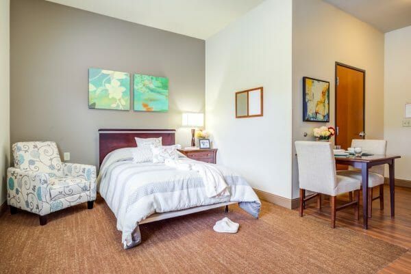 An assisted living studio apartment at Arlington Place Health Campus, furnished with a bed, an easy chair and a small dining table set for two