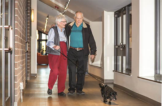 Kavod Senior Life hallway in the community with two residents/seniors walking a little dog