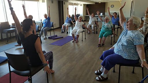 Kavod Senior Life yoga studio with teacher at the front and seniors sitting in chairs doing yoga poses and purple mats